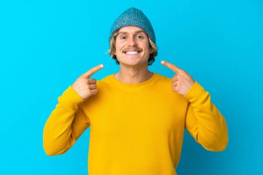 Mavi arka planda izole edilmiş yakışıklı sarışın adam baş parmak işareti yapıyor.