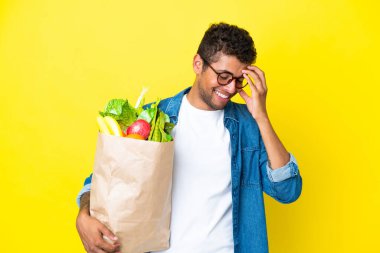 Sarı arka planda izole edilmiş alışveriş torbası tutan genç Brezilyalı adam gülüyor.