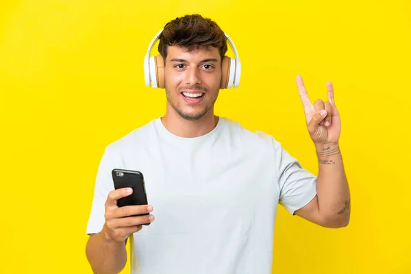 年轻的高加索帅哥 背景是黄色的 一边听着音乐 一边做着一个移动的摇滚手势 — 图库照片