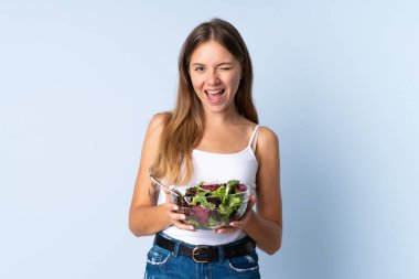 Genç Litvanyalı bir kadın mavi arka planda bir kase salata tutarken göz kırpıyor.