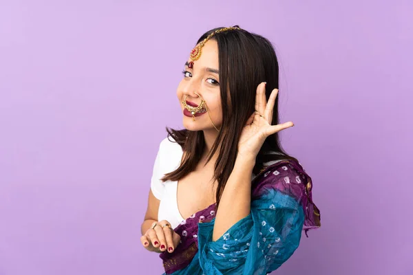 耳に手を置くことによって何かを聞く紫色の背景に隔離された若いインド人女性 — ストック写真