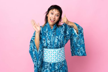 Kimono giyen genç bir kadın izole edilmiş mavi arka planda telefon jestleri yapıyor ve şüphe duyuyor.