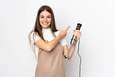 El blenderi kullanan genç bir kadın beyaz arka planda izole edilmiş, parmağını yana doğru gösteriyor.