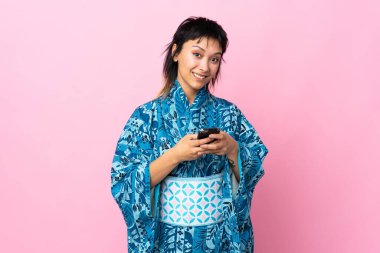 Kimono giyen genç bir kadın izole edilmiş mavi arka planda cep telefonuyla bir mesaj gönderiyor.