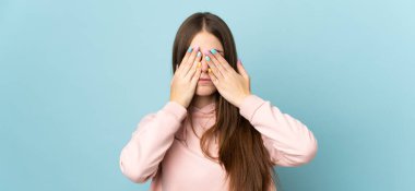 Mavi arka planda izole edilmiş genç beyaz kadın gözlerini elleriyle kapatıyor.