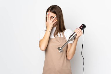 El blenderi kullanan genç bir kadın beyaz arka planda izole edilmiş gözleri kapatıyor ve parmaklarından bakıyor.