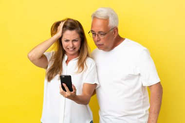 Sarı arka planda izole edilmiş orta yaşlı çift cep telefonuyla bir mesaj okuyor.