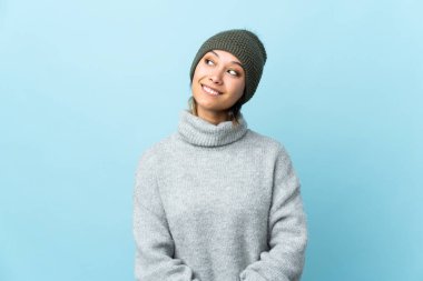 Kış şapkalı genç Uruguaylı kız mavi arka planda izole edilmiş gülüyor ve yukarı bakıyordu.