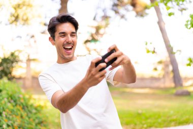 Parktaki genç beyaz adam cep telefonuyla oynuyor.