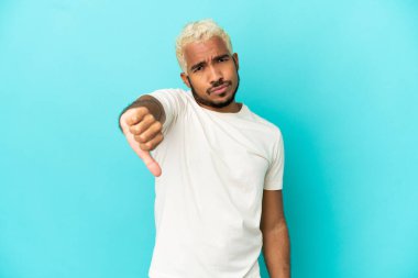Mavi arka planda izole edilmiş genç Kolombiyalı yakışıklı adam olumsuz ifadelerle baş parmağını aşağı gösteriyor.