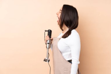 Genç esmer kız arka planda el blenderi kullanıyor. Yan pozisyonda gülüyor.