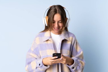 Genç Litvanyalı kadın mavi arka planda izole edilmiş müzik dinliyor ve cep telefonu arıyor.