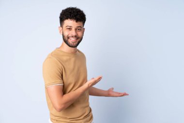 Faslı genç bir adam mavi arka planda izole edilmiş. Davet etmek için ellerini yana uzatıyor.