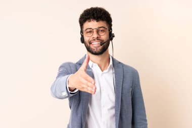 Faslı bir pazarlamacı bej arka planda izole edilmiş bir kulaklıkla çalışıyor. İyi bir anlaşma için el sıkışıyor.