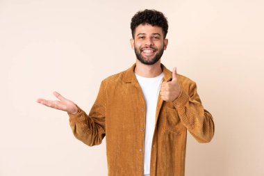 Faslı genç bir adam bej arka planda izole edilmiş. Kopyalama alanı hayali bir şekilde avucunun içinde duruyor. Bir ilan vermek için.