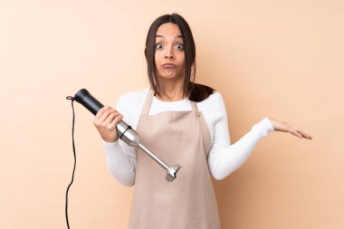 El blenderi kullanan genç esmer kız izole edilmiş arka planda şüpheleri var ve ellerini kaldırıyor.