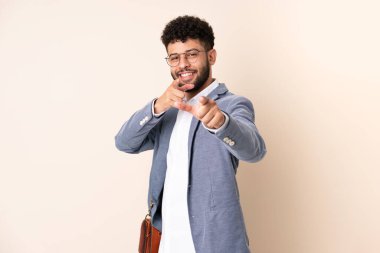 Faslı genç iş adamı bej arka planda izole edilmiş ön tarafı işaret ediyor ve gülümsüyor.