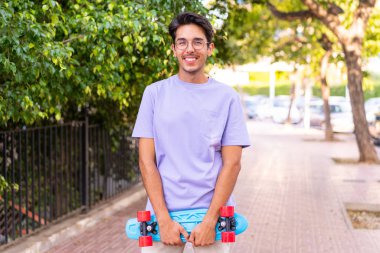 Parkta yüzünde mutlu bir ifadeyle paten yapan beyaz bir genç.