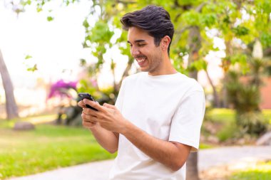 Parktaki genç beyaz adam cep telefonuyla mesaj ya da e-posta yolluyor.