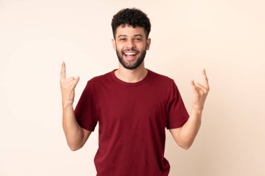 Bej arka planda izole edilmiş genç Faslı adam boynuz hareketi yapıyor.