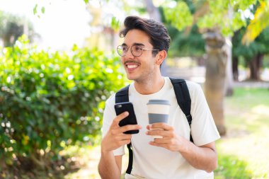 Dışarıda cep telefonu kullanan ve elinde kahve tutan genç bir öğrenci.