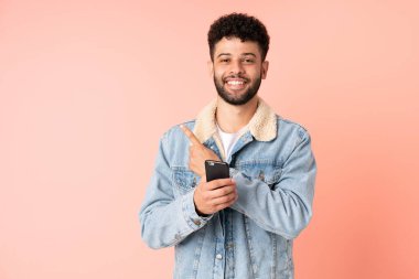 Pembe arka planda izole edilmiş cep telefonu kullanan genç Faslı bir adam bir ürünü sunmak için kenarı işaret ediyor.