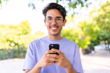Parkta genç beyaz bir adam cep telefonuyla mesaj yolluyor.
