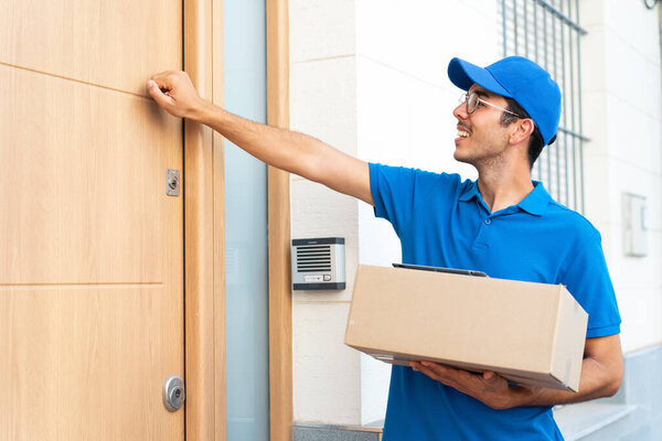 Young delivery man at outdoors holding boxes and knocking on the door