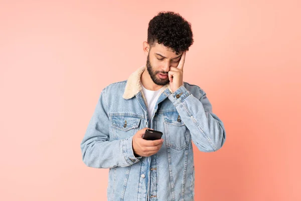 Young Moroccan man using mobile phone isolated on pink background with headache