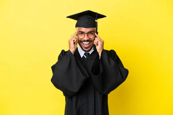 Joven Graduado Universitario Colombiano Aislado Sobre Fondo Amarillo Con Gafas —  Fotos de Stock