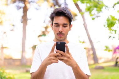 Parktaki genç beyaz adam cep telefonu kullanıyor.