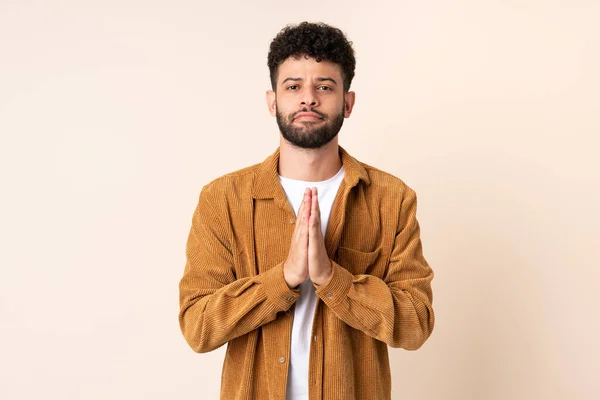 Young Moroccan man isolated on beige background keeps palm together. Person asks for something