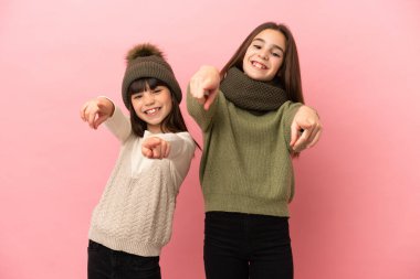 Pembe arka planda izole edilmiş kışlık giysiler giyen küçük kız kardeşler gülümserken parmaklarını sana gösteriyor.