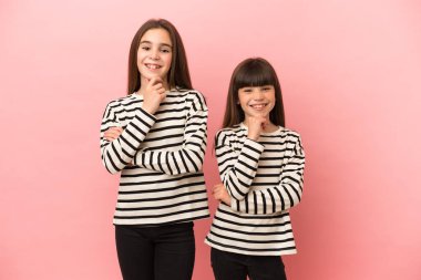 Küçük kız kardeşler pembe arka planda izole edilmiş gülümseyen ve kendinden emin bir yüzle öne bakan kızlar.