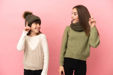 Pembe arka planda izole edilmiş kışlık giysiler giyen küçük kız kardeşler, parmak kaldırarak çözümü bulmaya çalışıyorlar.
