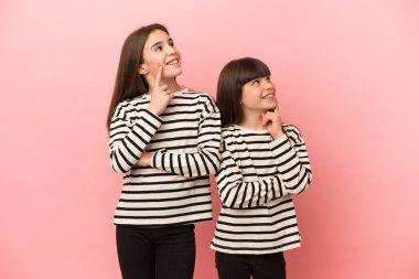 Küçük kız kardeşler pembe arka planda izole edilmiş bir şekilde yukarı bakarken bir fikir düşünüyorlar.