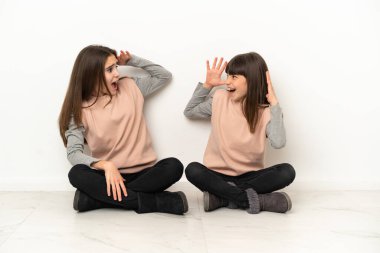 Beyaz arka planda izole edilmiş yerde oturan küçük kız kardeşler şaşırmış ve şok olmuş bir yüz ifadesiyle