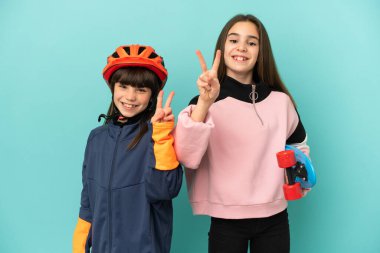 Küçük kız kardeşler bisiklet çalışıyor ve kaykaycı mavi arka planda izole edilmiş gülümsüyor ve zafer işareti gösteriyor.