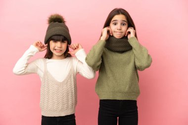 Pembe arka planda izole edilmiş kışlık giysiler giyen küçük kız kardeşler iki kulağını da elleriyle kapatıyor.