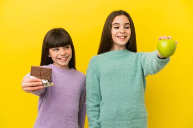 Sarı arka planda izole edilmiş küçük kız kardeşler bir elinde çikolata tableti, diğerinde elma alıyorlar.