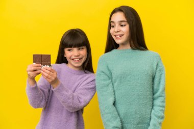 Sarı arka planda izole edilmiş küçük kız kardeşler çikolata tableti alıyor ve mutlular.