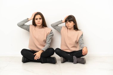 Beyaz arka planda izole bir şekilde yerde oturan küçük kız kardeşler hayal kırıklıklarıyla dolu ve anlamıyorlar.