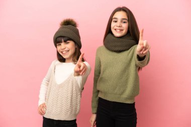 Pembe arka planda izole edilmiş kışlık giysiler giyen küçük kız kardeşler parmak kaldırıyor.