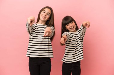 Pembe arka planda izole edilmiş küçük kız kardeşler sana gülümserken parmak atıyor.