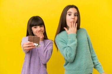 Sarı arka planda izole edilmiş küçük kız kardeşler çikolata tableti alıyor ve şaşırıyorlar.