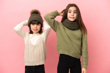 Pembe arka planda izole edilmiş kışlık giysiler giyen küçük kız kardeşler migreni olduğu için el ele tutuşuyorlar.