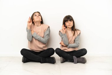 Küçük kız kardeşler beyaz arka planda izole bir şekilde yerde oturuyor parmaklarını çapraz yapıp en iyisini diliyorlar.