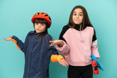 Küçük kız kardeşler bisiklet çalışıyor ve kaykaycı mavi arka planda izole edilmiş ellerini ve omuzlarını kaldırırken kuşkuları var.