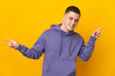 Sarı arka planda izole edilmiş genç beyaz adam parmak uçlarını işaret ediyor ve mutlu.