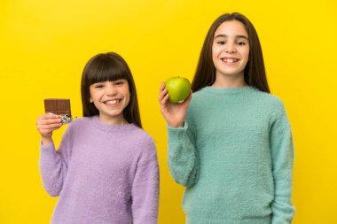 Sarı arka planda izole edilmiş küçük kız kardeşler bir elinde çikolata tableti, diğerinde elma alıyorlar.